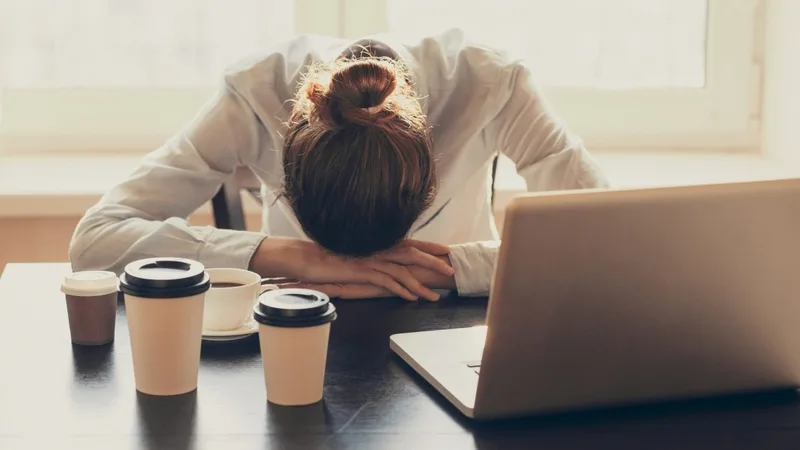 An office worker slumped over the desk beside a laptop and empty coffee cups — exhausted after hours of continuous computer work